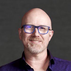 Photo if Ian Walker. A smiling white man, bald, wearing a blue shirt and blue glasses.